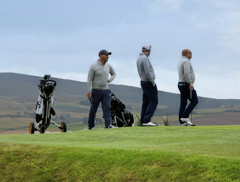 Three men in golf attire on a green course with rolling hills behind